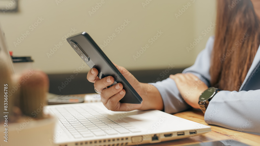 Fototapeta premium A woman is holding a cell phone while sitting at a desk with a laptop. She is focused on her phone, possibly checking messages or browsing the internet. The scene suggests a typical workday
