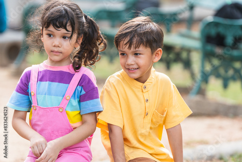 Two of multiracial little children smiling, sitting together outdoors in summer park. Adorable kids have fun playing in playground with active activity, holidays and vacations