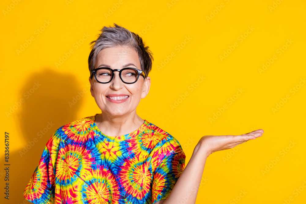 Photo of nice lovely retired woman wear stylish print clothes hold empty space isolated on yellow color background Photo of nice lovely retired woman wear stylish print clothes hold empty space isolated on yellow color background