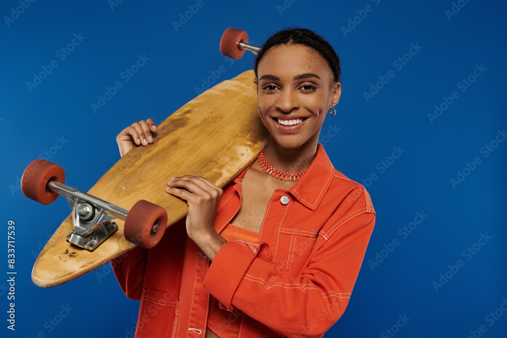 A smiling young African American woman in a vibrant orange outfit holds a skateboard against a blue background. A smiling young African American woman in a vibrant orange outfit holds a skateboard against a blue background.