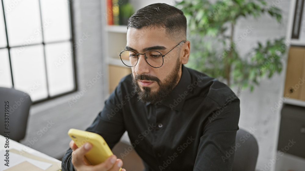 A focused young man with a beard and glasses using a smartphone in a modern office setting. A focused young man with a beard and glasses using a smartphone in a modern office setting.