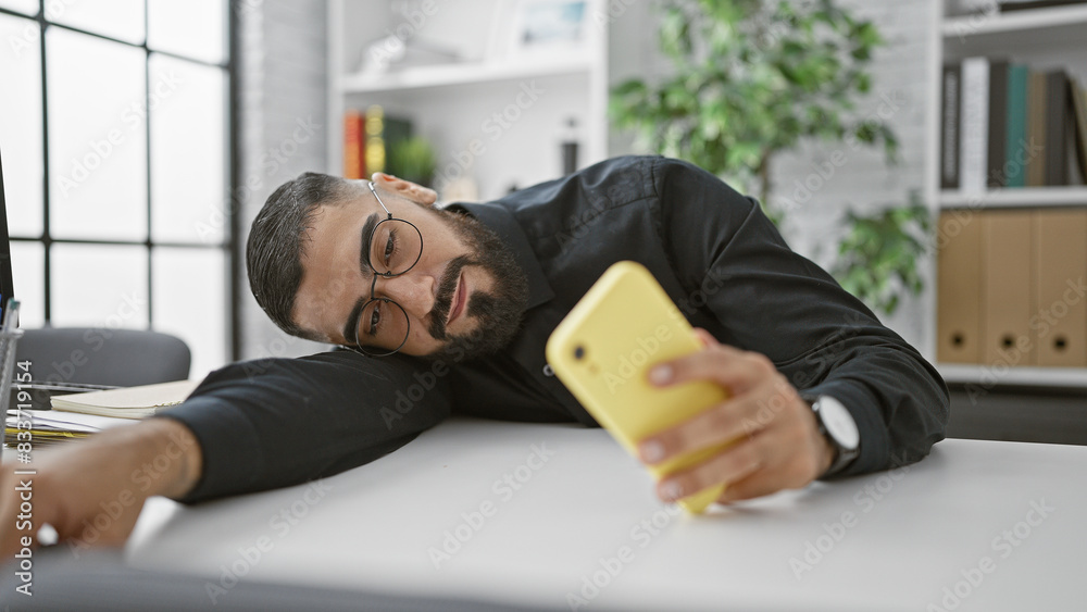 Obraz premium Tired young man with beard using smartphone at office desk indoors