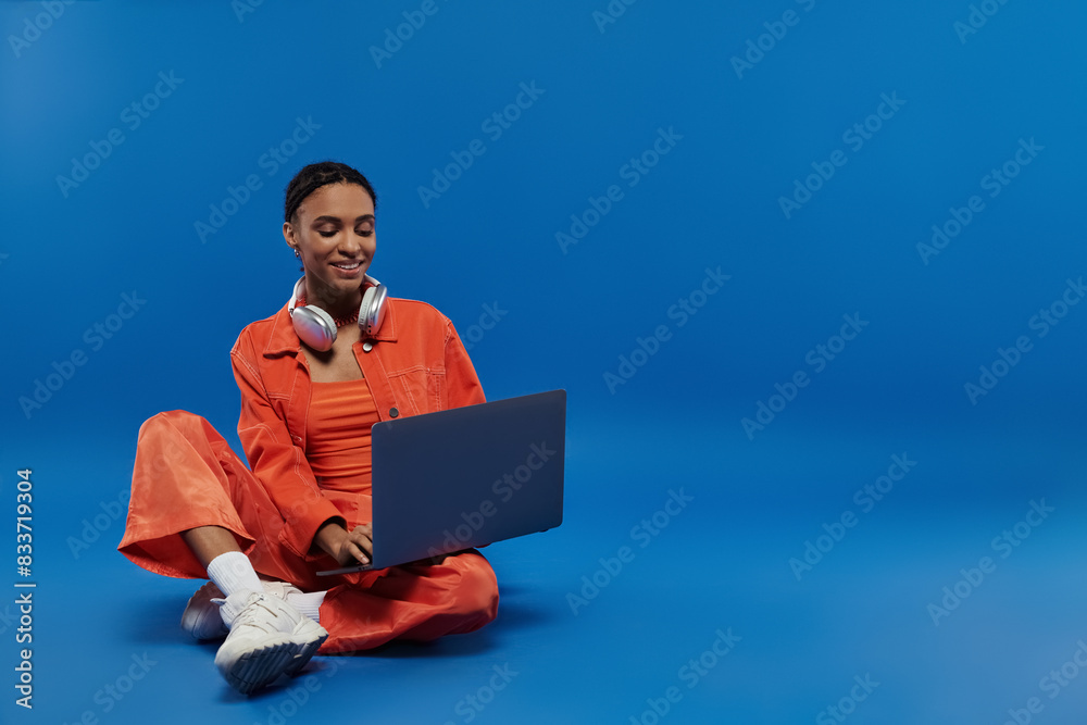 A young African American woman in a vibrant orange outfit sits on the floor with a laptop in front of her. A young African American woman in a vibrant orange outfit sits on the floor with a laptop in front of her.