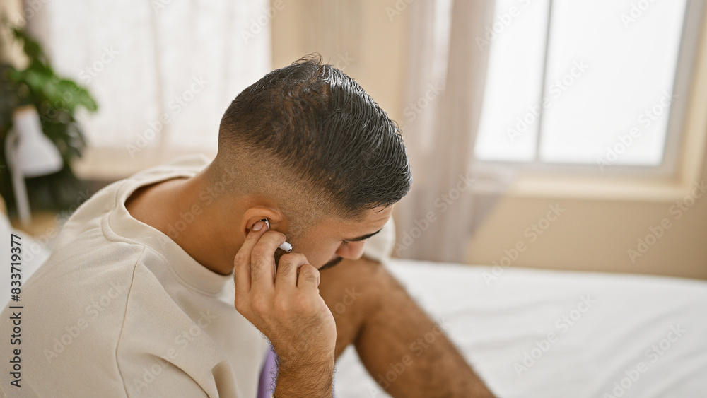 A thoughtful man with a beard in a white shirt touches a wireless earbud in a bright bedroom setting. A thoughtful man with a beard in a white shirt touches a wireless earbud in a bright bedroom setting.