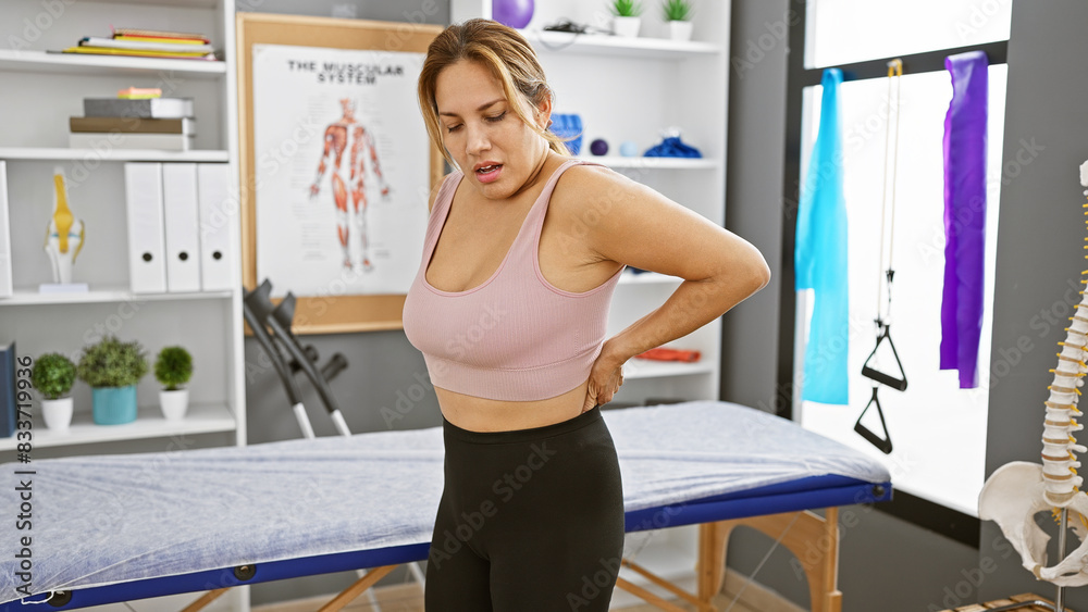 A young hispanic woman exhibits back pain inside a well-equipped rehabilitation clinic. A young hispanic woman exhibits back pain inside a well-equipped rehabilitation clinic.
