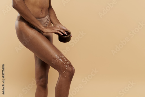 A stylish African American woman with a slim figure poses with coffee scrub on legs on a beige background. photo