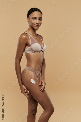 Pretty, slim African American woman poses confidently in bikini on beige background.