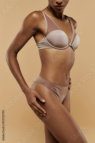 African American woman in bikini poses gracefully on beige background.