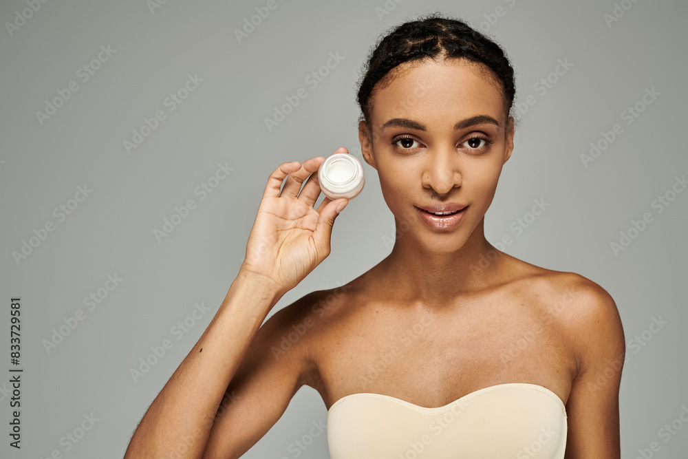 Young African American woman in strapless top, gracefully holds jar of cream on grey background. Young African American woman in strapless top, gracefully holds jar of cream on grey background.
