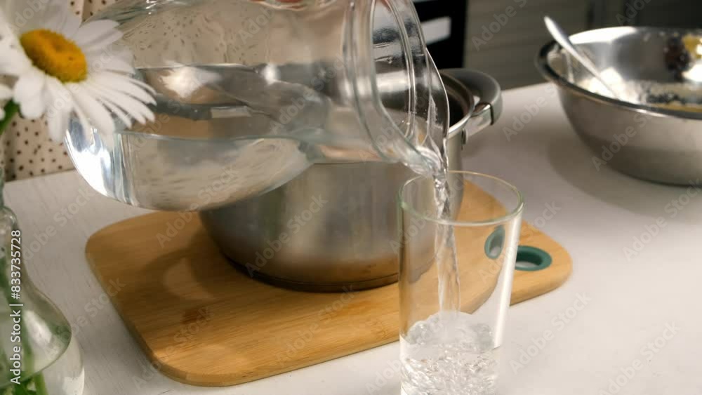 Woman pouring half of water glass to pan cooking on kitchen at home. Culinary cuisine, healthy eating, baking concept. Beautiful chamomile flowers in a vase at table.