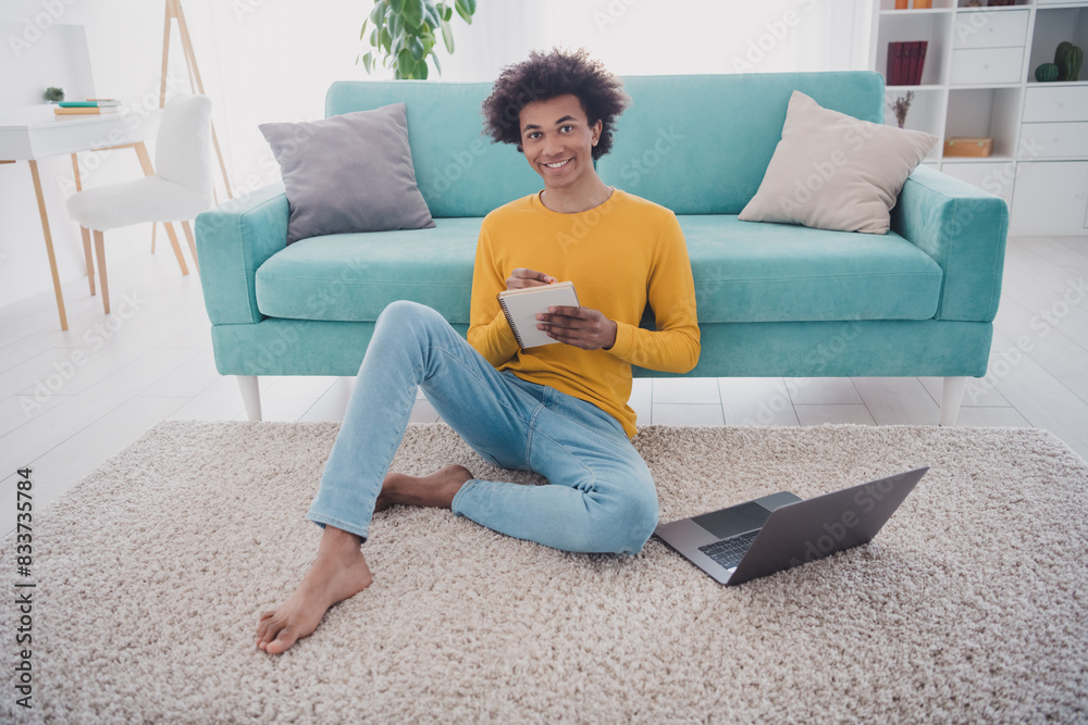Full length portrait of nice young man sit carpet laptop notepad wear pullover white interior flat indoors Full length portrait of nice young man sit carpet laptop notepad wear pullover white interior flat indoors