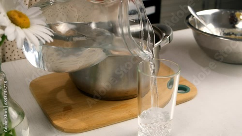 Woman pouring half of water glass to pan cooking on kitchen at home. Culinary cuisine, healthy eating, baking concept. Beautiful chamomile flowers in a vase at table.