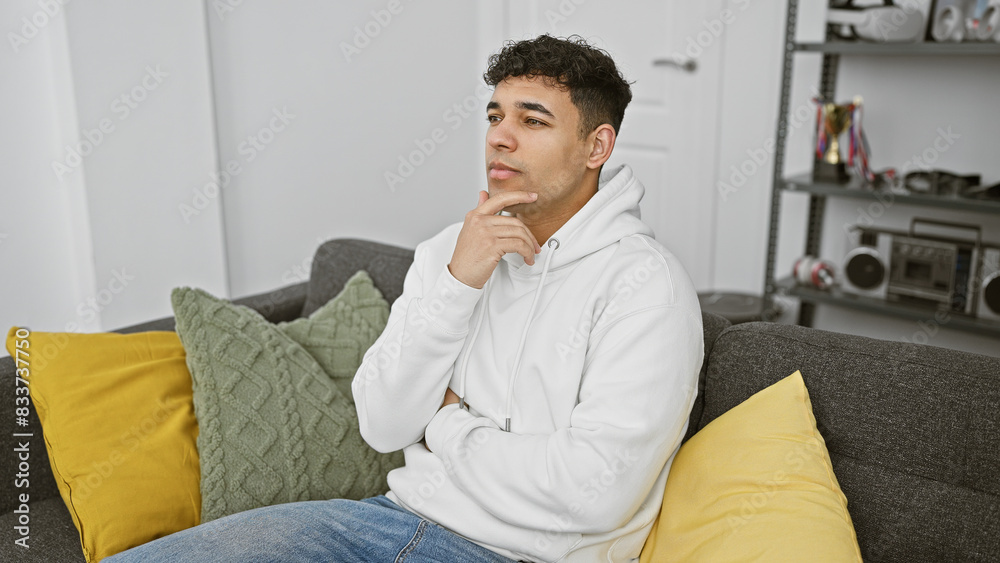 Pensive young man with beard in hoodie sitting on sofa in modern living room interior
