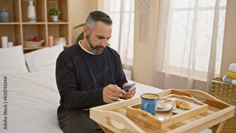 A mature hispanic man with grey beard using a smartphone in a well-lit bedroom, sitting by a breakfast tray. A mature hispanic man with grey beard using a smartphone in a well-lit bedroom, sitting by a breakfast tray.