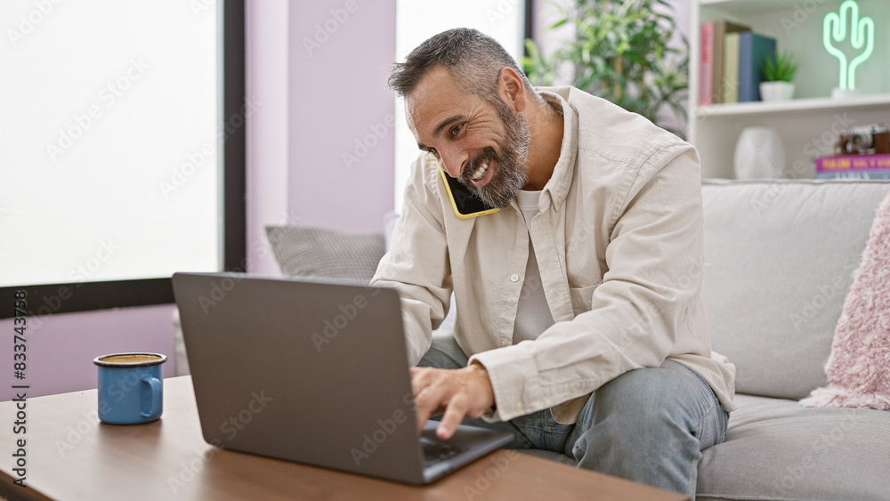 Smiling mature man with grey beard multitasking with phone and laptop in a cozy living room. Smiling mature man with grey beard multitasking with phone and laptop in a cozy living room.