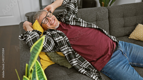 A content middle-aged man with grey hair, glasses, and casual clothes reclines on a sofa in a modern living room. photo