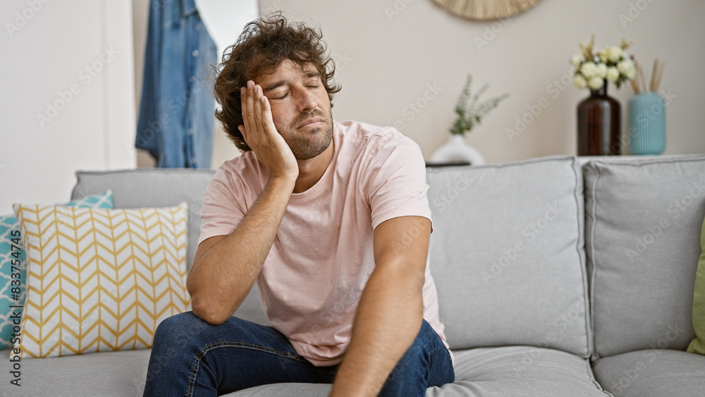 Tired young hispanic man seated on a gray couch in a well-decorated living room, exuding a casual, domestic vibe. Tired young hispanic man seated on a gray couch in a well-decorated living room, exuding a casual, domestic vibe.