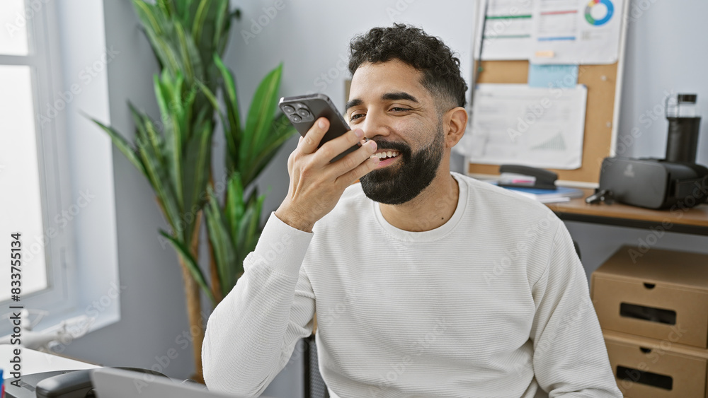 A smiling young man with a beard in an office setting talks on a smartphone, conveying a casual professional vibe. A smiling young man with a beard in an office setting talks on a smartphone, conveying a casual professional vibe.