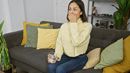 Surprised latina woman in yellow sweater holding a glass of water in a cozy living room
