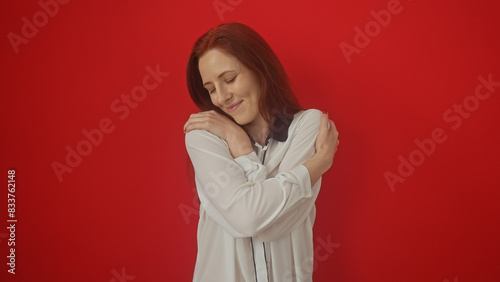 Serene redhead woman embracing herself against a vibrant red isolated background expressing self-love and confidence.