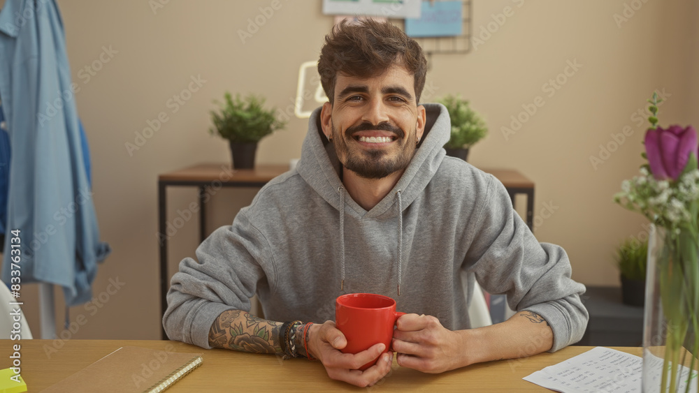 A cheerful young hispanic man with a beard and tattoos holds a red mug, sitting at a table indoors in a cozy apartment setting. A cheerful young hispanic man with a beard and tattoos holds a red mug, sitting at a table indoors in a cozy apartment setting.