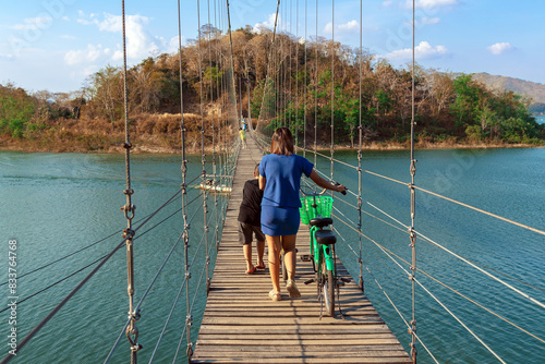 Back view of female tourist with son leads bicycle and enjoy nature across a high sling suspension bridge to the island with green trees. Family leads bicycle across wooden bridge over river to park.