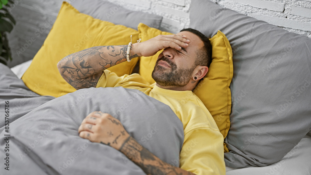 Fototapeta premium Tattooed hispanic man with beard in bedroom feeling headache or stress while lying in bed.