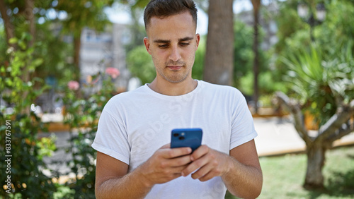 A handsome young man texts on his smartphone in a lush green city park wearing a casual white t-shirt.