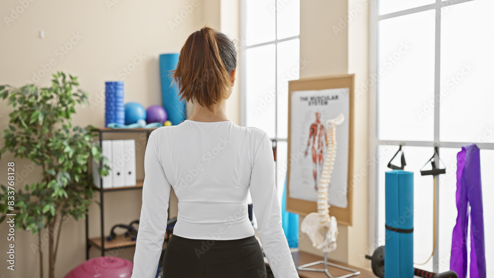A young adult woman stands in a gym, facing away, surrounded by fitness equipment and anatomy posters. A young adult woman stands in a gym, facing away, surrounded by fitness equipment and anatomy posters.