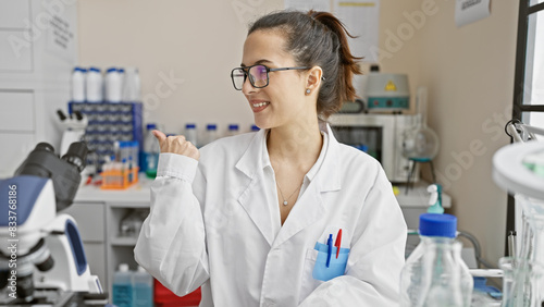 Smiling young hispanic woman scientist in lab coat pointing sideways in a laboratory setting.