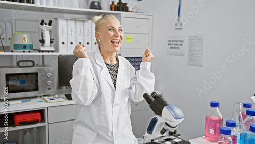 Attractive young blonde woman scientist celebrating a winning discovery  microscope analysis in a bustling lab brings a joyful smile 