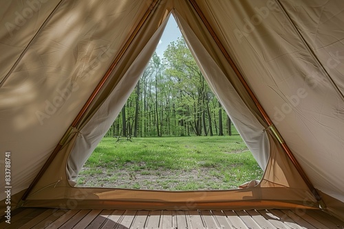 The view from inside an outdoor beige tent with canvas walls, wooden floors and roof, looking out onto green grass in springtime through its triangular opening. the outside is surrounded by trees.