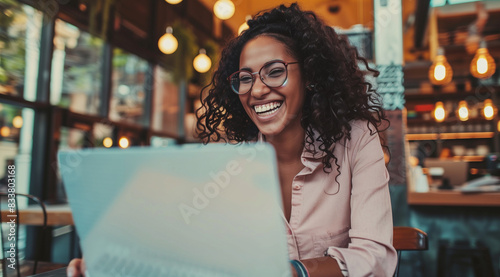 Happy young black woman working remotely on laptop in a cafe. African american remote worker laughing with colleagues on a virtual video call team meeting. Digital nomad freelancing  photo