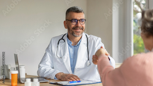 Smiling male doctor shaking hand with elderly female patient at consultation in hospital. Elderly woman thanking for therapy and support photo