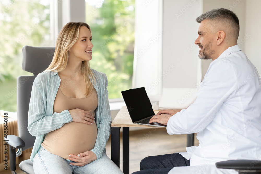Male gynecologist doctor using laptop computer and talking to pregnant European woman at hospital. Pregnancy, medicine, healthcare concept Male gynecologist doctor using laptop computer and talking to pregnant European woman at hospital. Pregnancy, medicine, healthcare concept