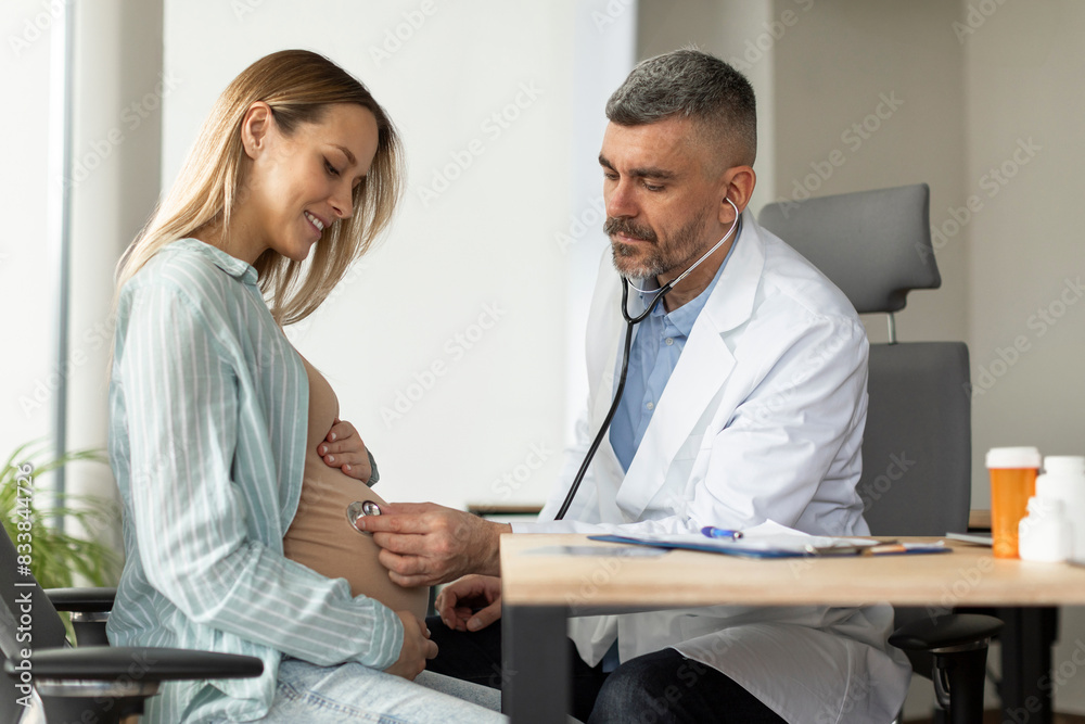 Obraz premium Pregnant woman having appointment with male doctor at clinic, gynecologist OB GYN medic specialist with stethoscope listens to baby's heartbeat in mother's belly