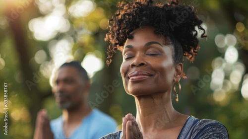A woman with curly hair wearing a blue top has her eyes closed and is smiling while a man in a blue shirt stands behind her with his hands together in a prayer position both in a peaceful outdoor