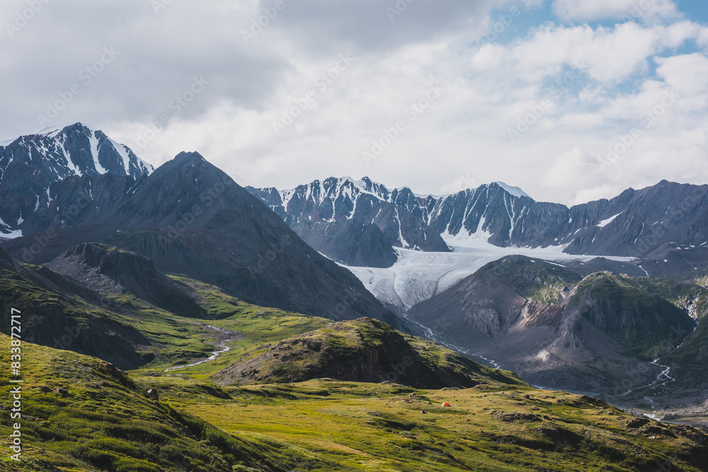 Dramatic scenery in alpine valley with creek among green hills and rocks with view to rocky sharp pointy peak, large snow-capped peaked top, mountain range and big glacier tongue under gray cloudy sky Dramatic scenery in alpine valley with creek among green hills and rocks with view to rocky sharp pointy peak, large snow-capped peaked top, mountain range and big glacier tongue under gray cloudy sky
