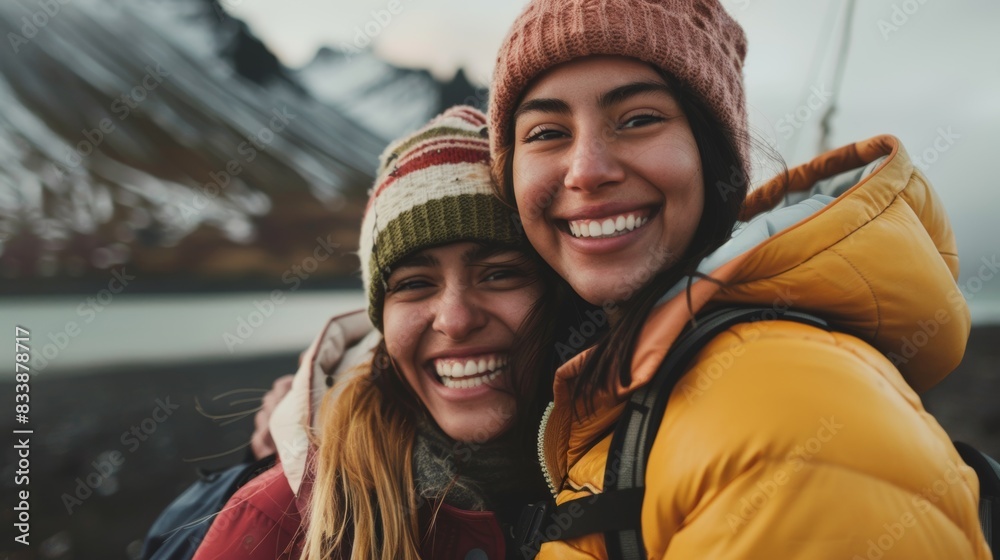Two smiling women in winter hats and jackets embracing each other in a mountainous landscape with a body of water in the background under a cloudy sky. Two smiling women in winter hats and jackets embracing each other in a mountainous landscape with a body of water in the background under a cloudy sky.
