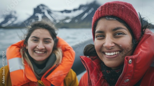 Two women smiling wearing life jackets and red hats standing on a boat with snow-capped mountains in the background.