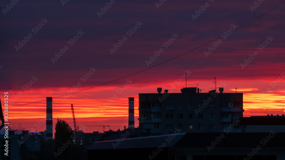 Urban Sunset. A dramatic sunset sky over an urban landscape with silhouettes of buildings and industrial elements against a vibrant backdrop of red and orange hues.