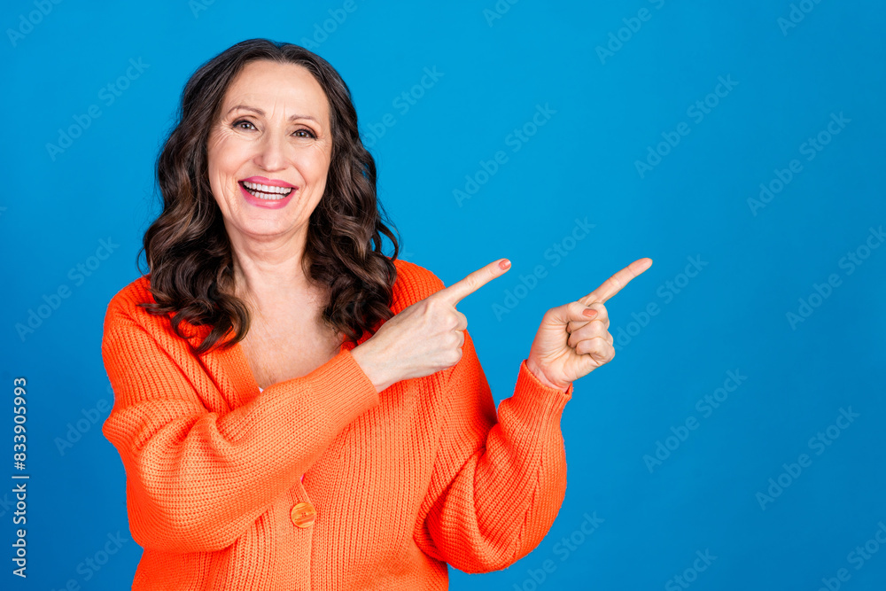 Photo portrait of gorgeous senior woman point empty space wear trendy orange outfit hairdo isolated on blue color background Photo portrait of gorgeous senior woman point empty space wear trendy orange outfit hairdo isolated on blue color background