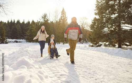 Parents take children on sleds while happily walking along snowy path in winter park. Man pulls sled with his son and daughter and smiling woman follows them. Concept of family winter fun.