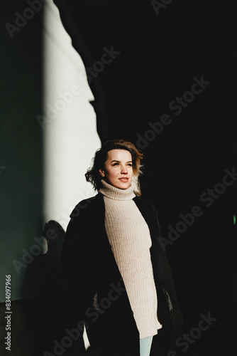 A woman in a black coat and white sweater stands against a wall, smiling warmly at the camera. The image captures a moment of joy and simplicity, perfect for lifestyle and fashion concepts.