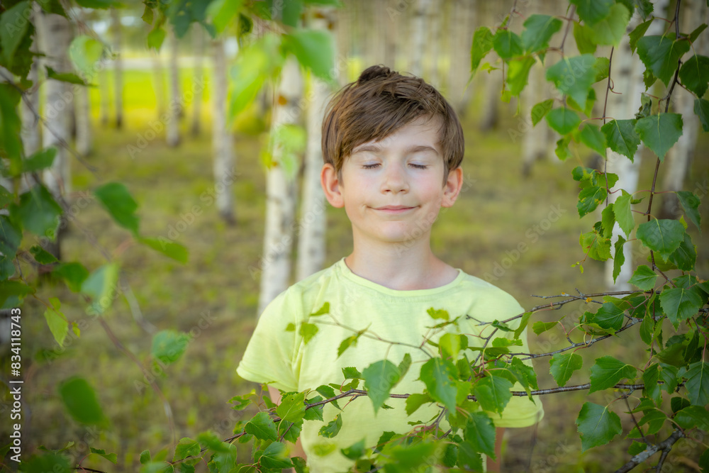 A young boy wearing a light green shirt smiles while holding a leafy branch in a birch tree forest. The background features rows of birch trees, creating a serene and natural setting. A young boy wearing a light green shirt smiles while holding a leafy branch in a birch tree forest. The background features rows of birch trees, creating a serene and natural setting.