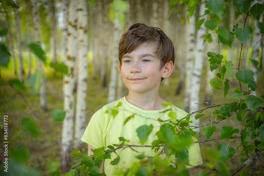A young boy wearing a light green shirt smiles while holding a leafy branch in a birch tree forest. The background features rows of birch trees, creating a serene and natural setting. A young boy wearing a light green shirt smiles while holding a leafy branch in a birch tree forest. The background features rows of birch trees, creating a serene and natural setting.