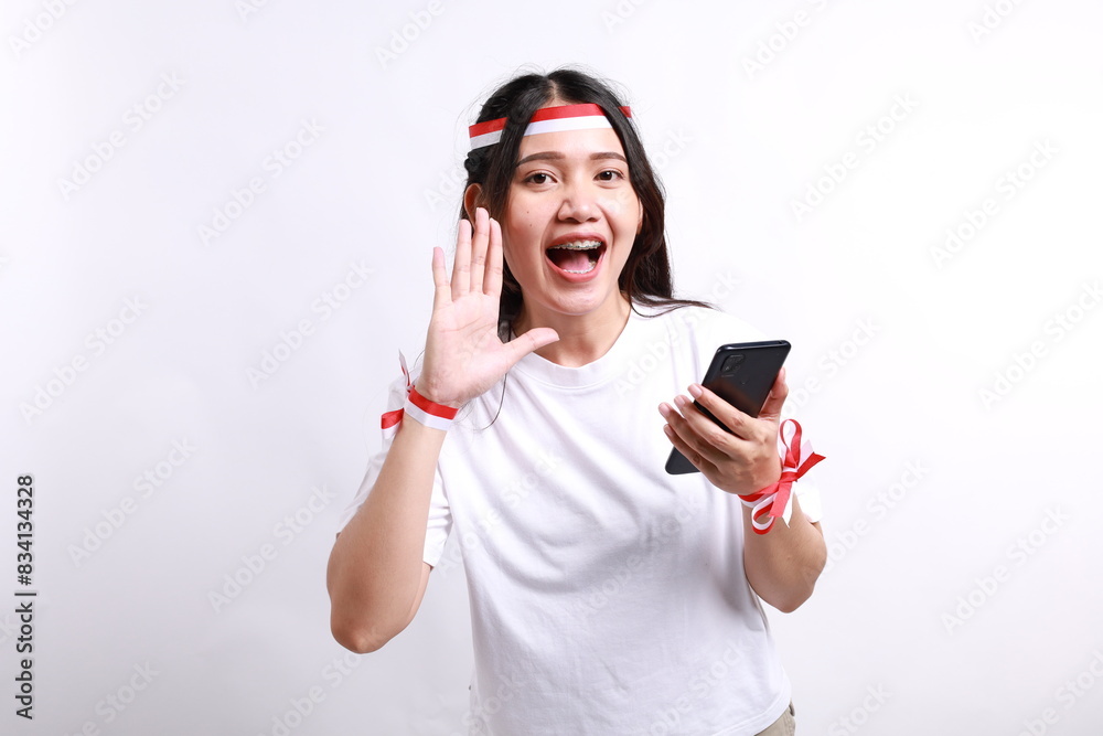 Naklejka premium Young beautiful woman wearing red white flag ribbon holding her phone while shouting and screaming loud with a hand on her mouth. Indonesia's independence day concept.