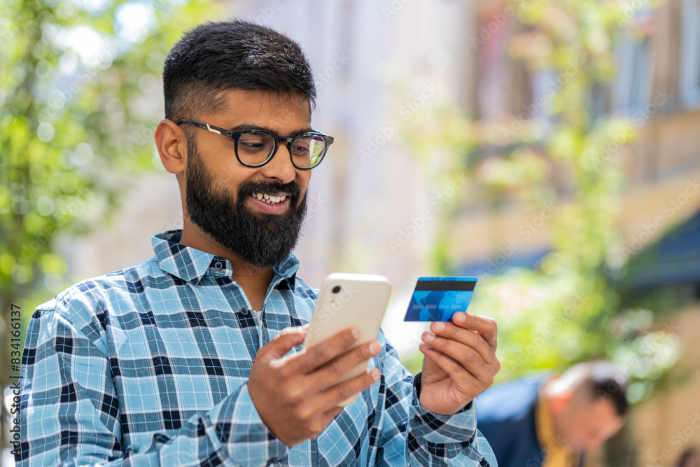 Indian bearded man using credit bank card smartphone while transferring money, purchases online shopping, order food delivery booking hotel room outdoors. Hispanic guy on urban city street. Horizontal Indian bearded man using credit bank card smartphone while transferring money, purchases online shopping, order food delivery booking hotel room outdoors. Hispanic guy on urban city street. Horizontal