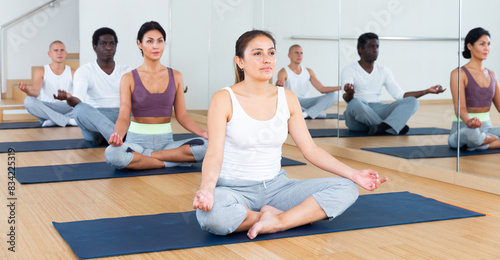 Concentrated young Latina relaxing and meditating in lotus pose with group of people in fitness studio