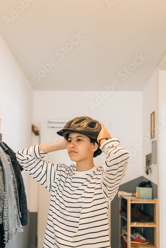 A teenage is putting on a helmet, she is wearing a striped shirt and is standing in a hallway